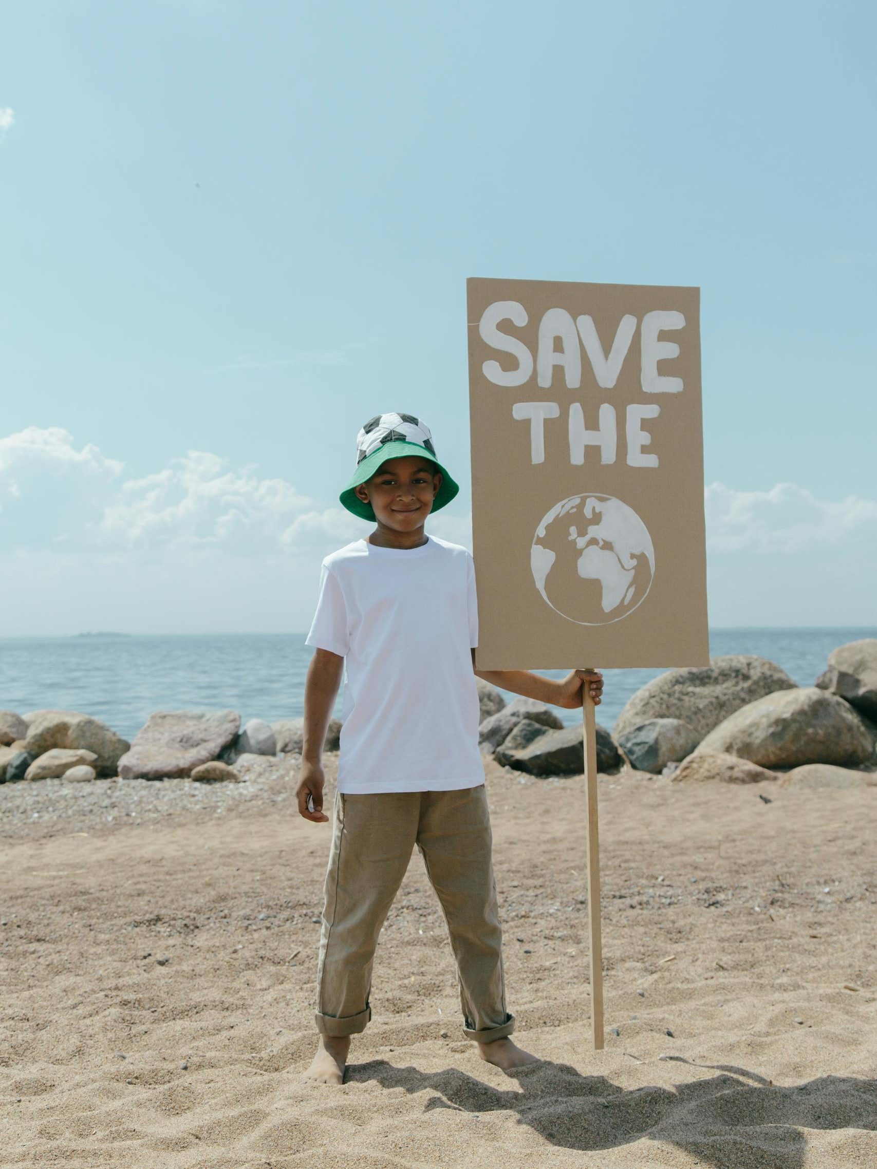 Young boy holding a Save the Earth sign on a sandy beach with rocks.